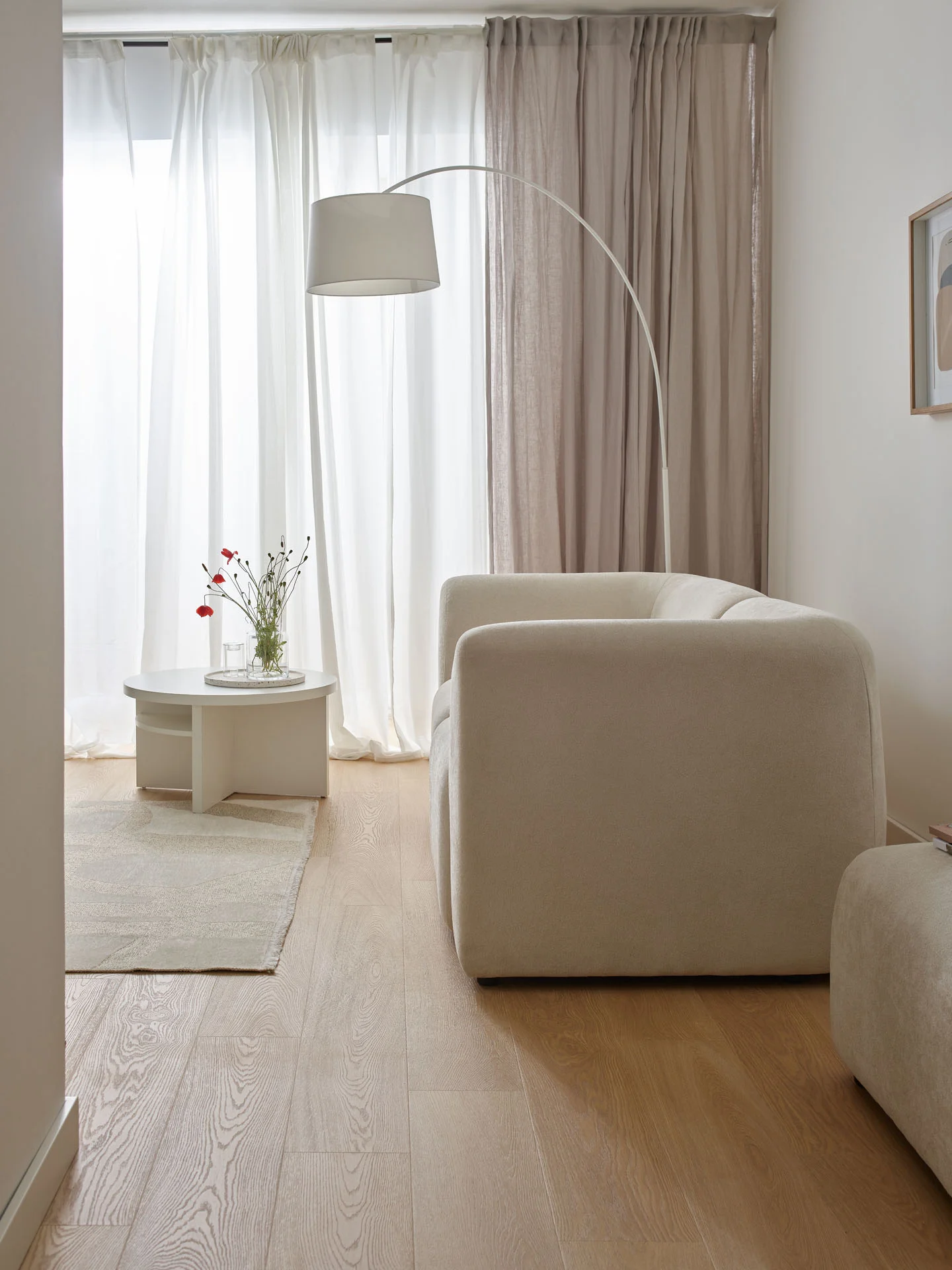 Biała sofa ustawiona obok dużych okien z jasnymi zasłonami, które wpuszczają naturalne światło i wizualnie podnoszą sufit.